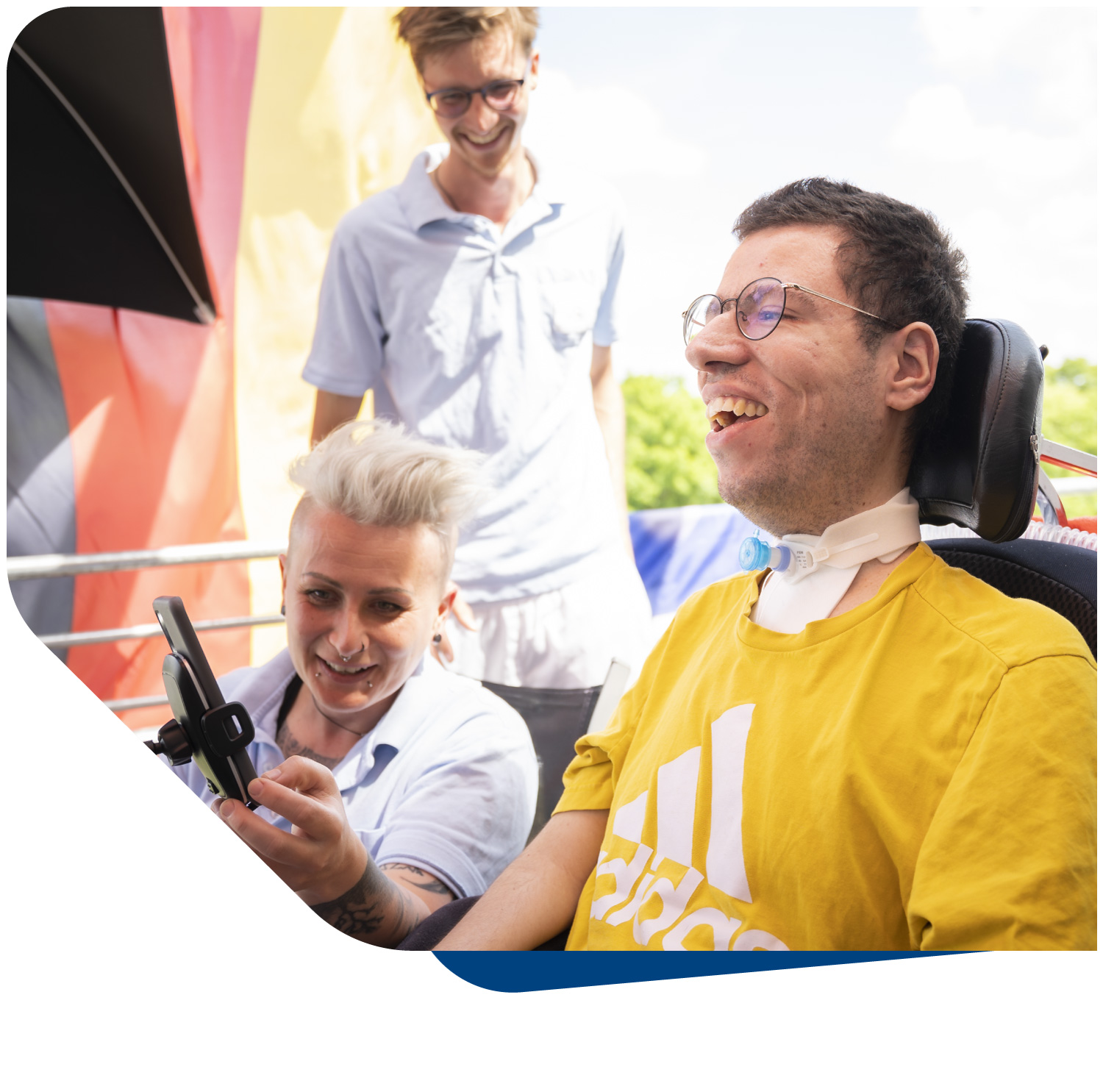 Eine fröhliche Szene im Freien, die einen lächelnden Patienten im Rollstuhl zeigt, der von zwei zugewandten männlichen Pflegeassistenten begleitet wird. Der Patient im gelben T-Shirt lacht ausgelassen und blickt nach oben, während ein Pflegeassistent ihm möglicherweise bei einer Aktivität assistiert. Der zweite Assistent steht im Hintergrund und lächelt ebenfalls. Das Bild verdeutlicht das Konzept der Pflege-Assistenz des Intensivpflegedienstes Kompass GmbH, das darauf abzielt, die Fähigkeiten der betreuten Menschen zu unterstützen und Freiräume zu ermöglichen, sodass Lebensqualität im Mittelpunkt des Handelns steht.