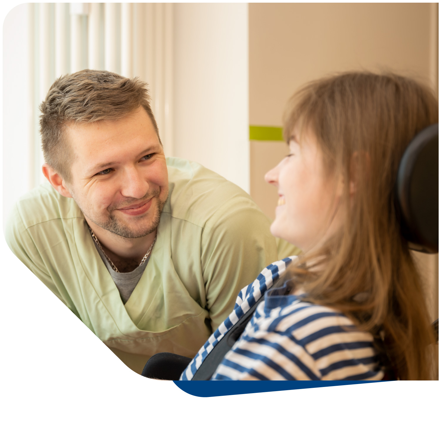 Ein freundlicher, lächelnder Pflegemitarbeiter blickt zugewandt eine sitzende Patientin an, die ebenfalls lächelt und in einem Rollstuhl oder speziellen Stuhl sitzt. Das Bild fängt einen warmen Moment des Dialogs und der Verbundenheit ein und symbolisiert die vertrauensvolle Beziehung zwischen Pflegepersonal und Patienten. Es unterstreicht das Engagement des Intensivpflegedienstes Kompass, kranke und behinderte Menschen in ihrem selbstbestimmten Leben im eigenen Zuhause oder in ambulant betreuten Wohngemeinschaften im Raum München zu unterstützen.