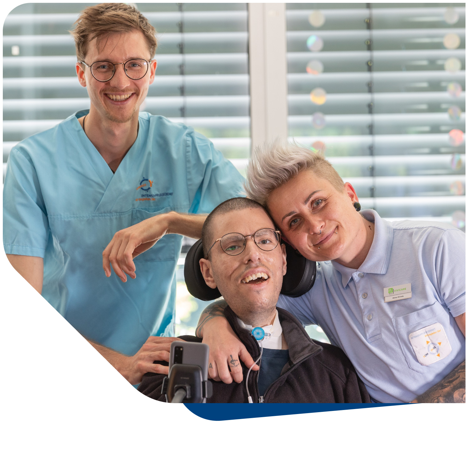 Eine herzliche Szene, die drei Personen zeigt, die alle freundlich in die Kamera lächeln. Im Vordergrund sitzt ein Patient im Rollstuhl mit einer Kanüle am Hals, der von zwei engagierten Pflegekräften, einer Frau mit kurzen, hellen Haaren und einem Mann in OP-Kleidung, umgeben ist. Die positive Ausstrahlung der Gruppe symbolisiert die patientenzentrierte Philosophie des Intensivpflegedienstes Kompass GmbH und das harmonische Miteinander, das im Pflegekonzept verankert ist. Das Bild vermittelt Empathie, Nähe und eine professionelle, zugleich persönliche Betreuung.