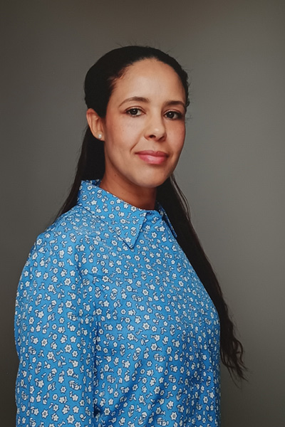 Porträt von Nadya Zouani, stellvertretende Pflegedienstleitung der KOMPASS GmbH München. Sie trägt eine blau-weiß gemusterte Bluse, hat langes dunkles Haar und blickt mit einem freundlichen, kompetenten Lächeln in die Kamera vor einem neutralen grauen Hintergrund.