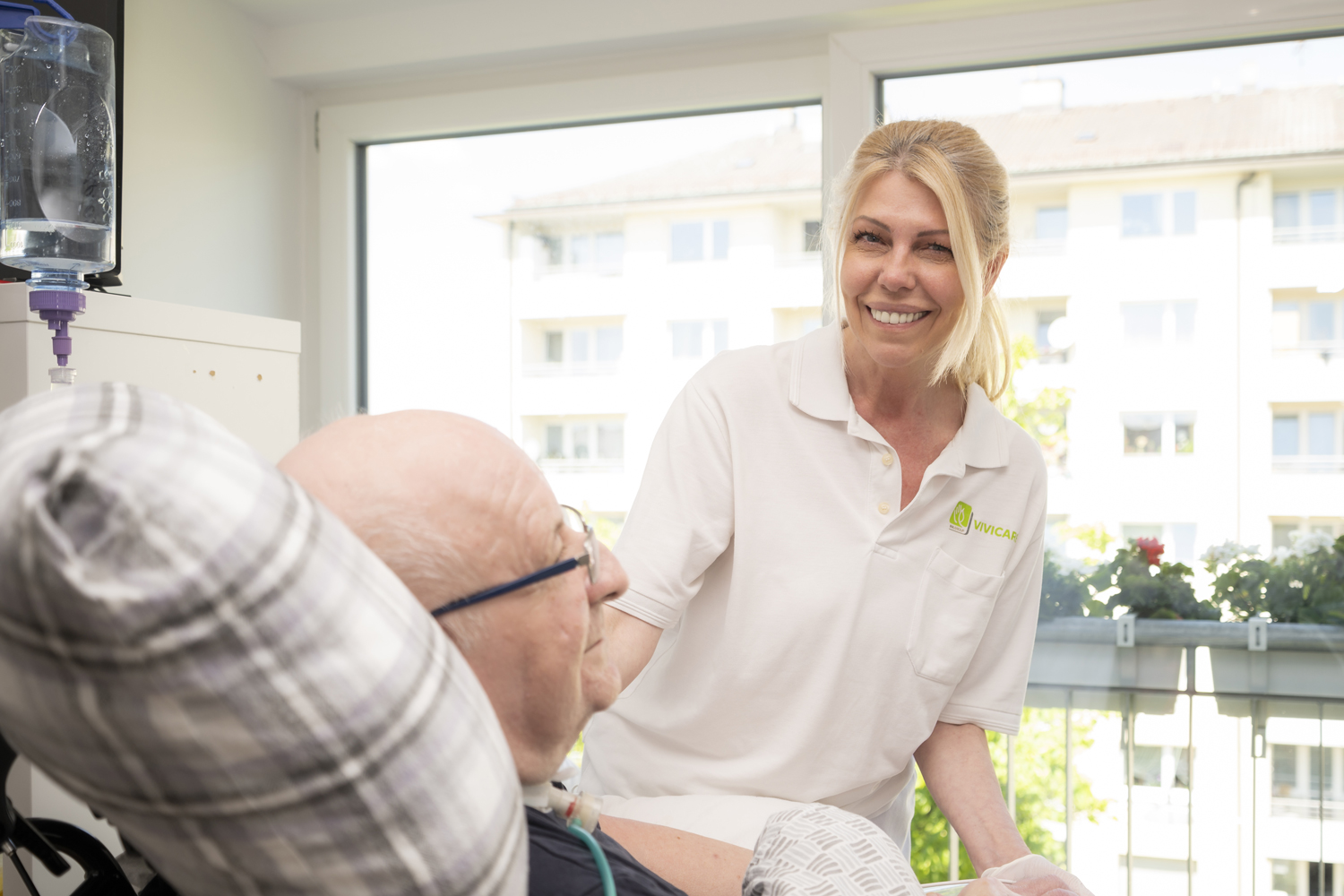 Eine lächelnde Pflegefachkraft mit blonden Haaren in einem weißen Poloshirt blickt den Betrachter freundlich an, während sie sich einem Patienten zuwendet, der bequem in einem Pflegebett oder -sessel liegt. Der Patient trägt eine Brille und ist teilnahmsvoll. Im Hintergrund ist ein Fenster zu sehen, das den Blick auf eine städtische Wohngegend mit einem Balkon und Pflanzen freigibt, was die häusliche Umgebung betont. Über dem Patienten ist medizinische Ausrüstung sichtbar, die auf eine notwendige Betreuung hindeutet. Das Bild veranschaulicht das Konzept des selbstbestimmten Lebens zuhause und die professionelle, aber persönliche Unterstützung, die der Intensivpflegedienst Kompass GmbH seinen Patienten in ihrer vertrauten Umgebung bietet.