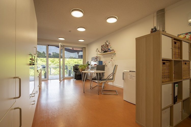 Ein heller und geräumiger Gemeinschaftsraum innerhalb der ambulanten Wohngemeinschaft in der Belgradstrasse 106 in München/Schwabing. Der Raum verfügt über Holzböden, große Fenster, die viel Tageslicht hereinlassen und den Blick auf Grünflächen freigeben. Eine Essecke mit Tisch und Stühlen sowie Regale sind sichtbar, was eine wohnliche und funktionale Atmosphäre für die Bewohner schafft.