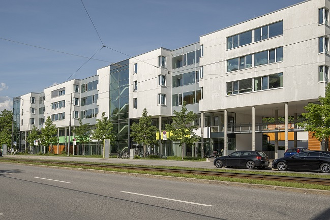 Die Außenansicht eines modernen, mehrstöckigen Wohngebäudes in der Belgradstrasse 106 in München/Schwabing. Die klare, weiße Architektur mit großen Fenstern und Balkonen vermittelt ein Gefühl von Offenheit und moderner Wohnqualität. Das Bild zeigt das Gebäude in einer urbanen Umgebung, umgeben von Bäumen und einer Straße.