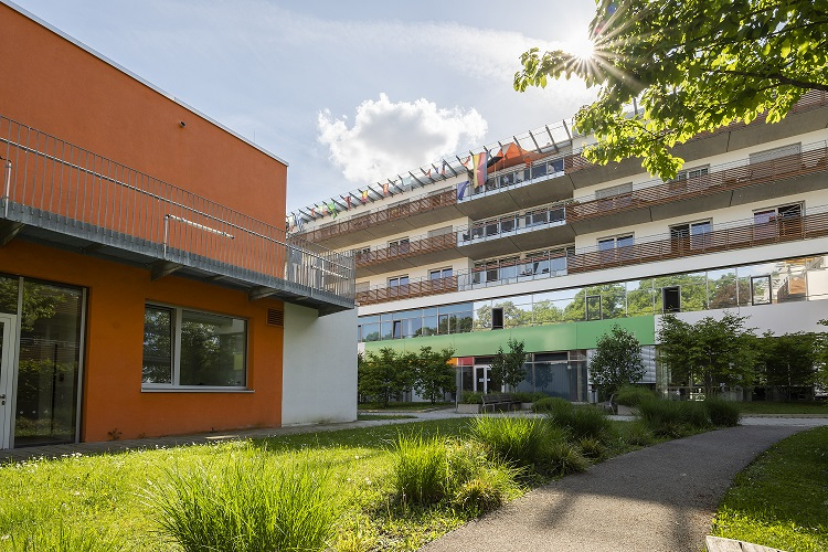 Eine Außenansicht des Gebäudekomplexes der ambulanten Wohngemeinschaft in der Belgradstrasse 106 in München/Schwabing. Das Bild zeigt einen Teil des Gebäudes mit einer rotbraunen Basis und einem modernen oberen Bereich, eingebettet in eine gepflegte Grünanlage mit Bäumen. Es unterstreicht die Kombination aus städtischer Lage und grüner Umgebung, nahe dem Luitpoldpark.