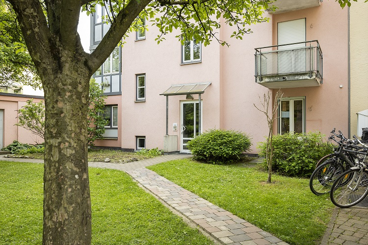Eine Außenansicht des Gebäudes der ambulanten Wohngemeinschaft in der Karl-Hromadnikstrasse in München/Pasing. Die Fassade des mehrstöckigen Gebäudes ist in einem hellen, freundlichen Farbton gehalten, mit vielen Fenstern, die auf eine lichtdurchflutete Inneneinrichtung schließen lassen. Das Bild zeigt das gepflegte Umfeld der Wohngemeinschaft.