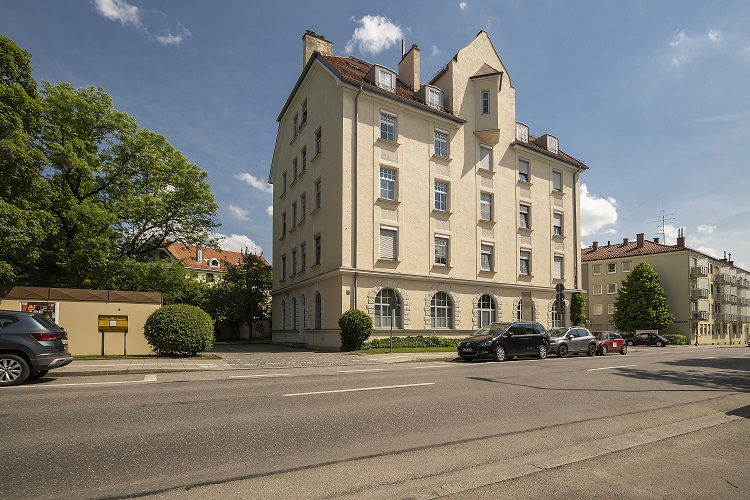 Eine repräsentative Außenansicht des Hauptgebäudes der ambulanten Wohngemeinschaft in der Karl-Hromadnikstrasse in München/Pasing. Das mehrstöckige, klassisch anmutende Gebäude ist von der Straße aus sichtbar, mit geparkten Fahrzeugen im Vordergrund, die eine zentrale Lage in einer städtischen Umgebung andeuten.